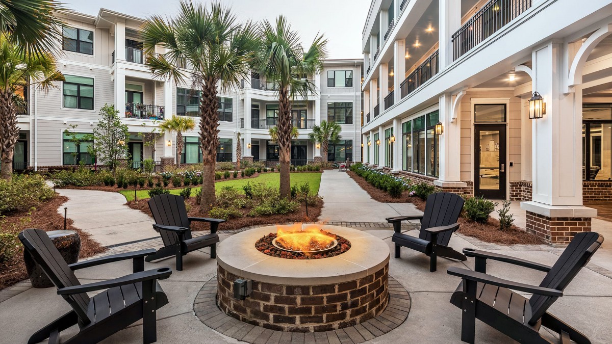 A modern apartment complex courtyard featuring a central fire pit surrounded by outdoor seating and landscaped with palm trees.