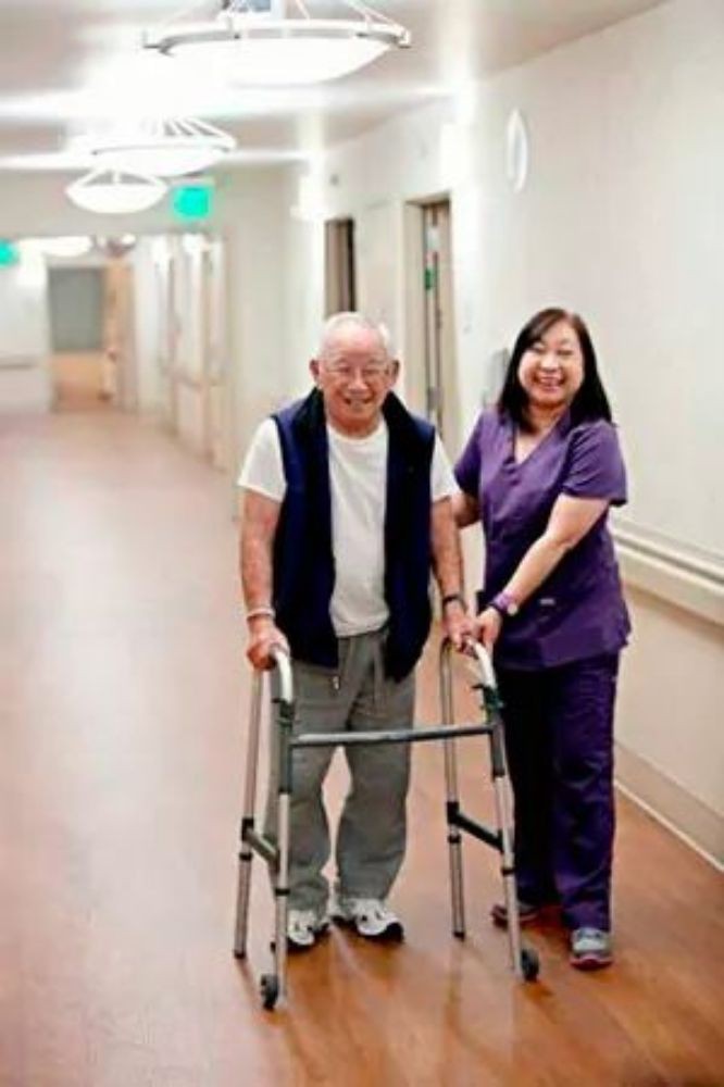 An elderly man using a walker is assisted by a nurse in a hospital corridor.