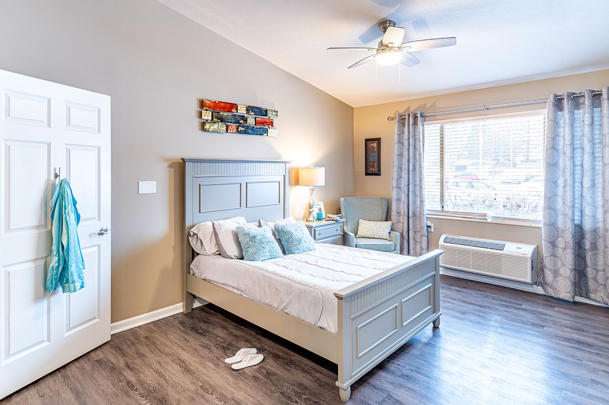 A cozy and well-decorated bedroom featuring a bed, lamp, artwork, and a large window allowing natural light to fill the space.