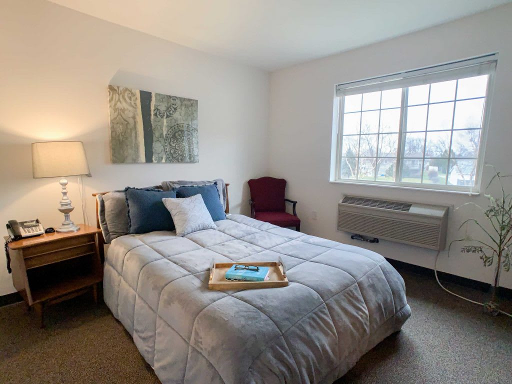 A cozy bedroom with a neatly made bed, artwork on the wall, and natural light streaming through the window.