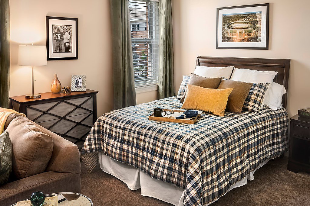 A cozy bedroom with plaid bedding and decorative pillows, featuring warm lighting and artwork on the walls.