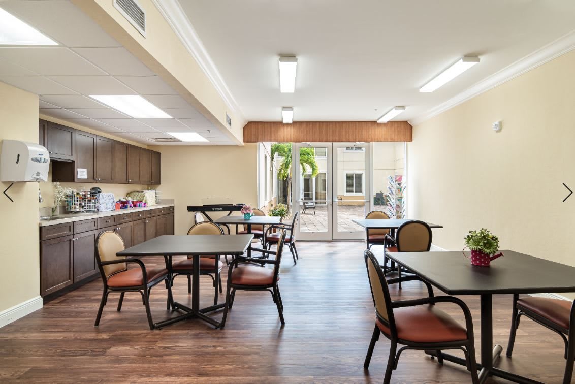 A well-lit dining area with multiple tables and chairs, featuring a kitchenette and large windows.