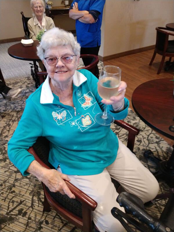 An elderly woman wearing glasses is smiling and sitting indoors, holding a glass in her hand.