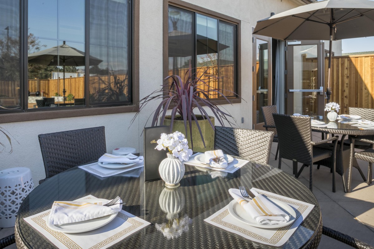A stylish outdoor patio dining area with a glass table set for four, featuring elegant place settings and decorative plants.