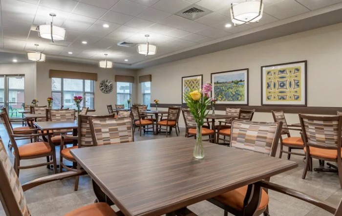 A cozy dining room with neatly arranged tables and chairs, decorated with flowers and artwork on the walls.