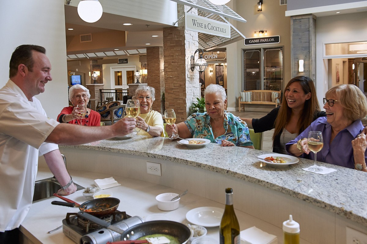 A group of seniors enjoying a meal and raising glasses in celebration at a kitchen counter.