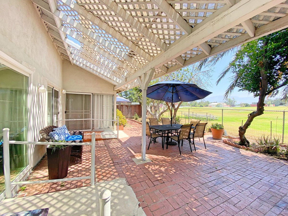 A sunny patio with a pergola, featuring a table and chairs under an umbrella, surrounded by plants and overlooking a fenced area.