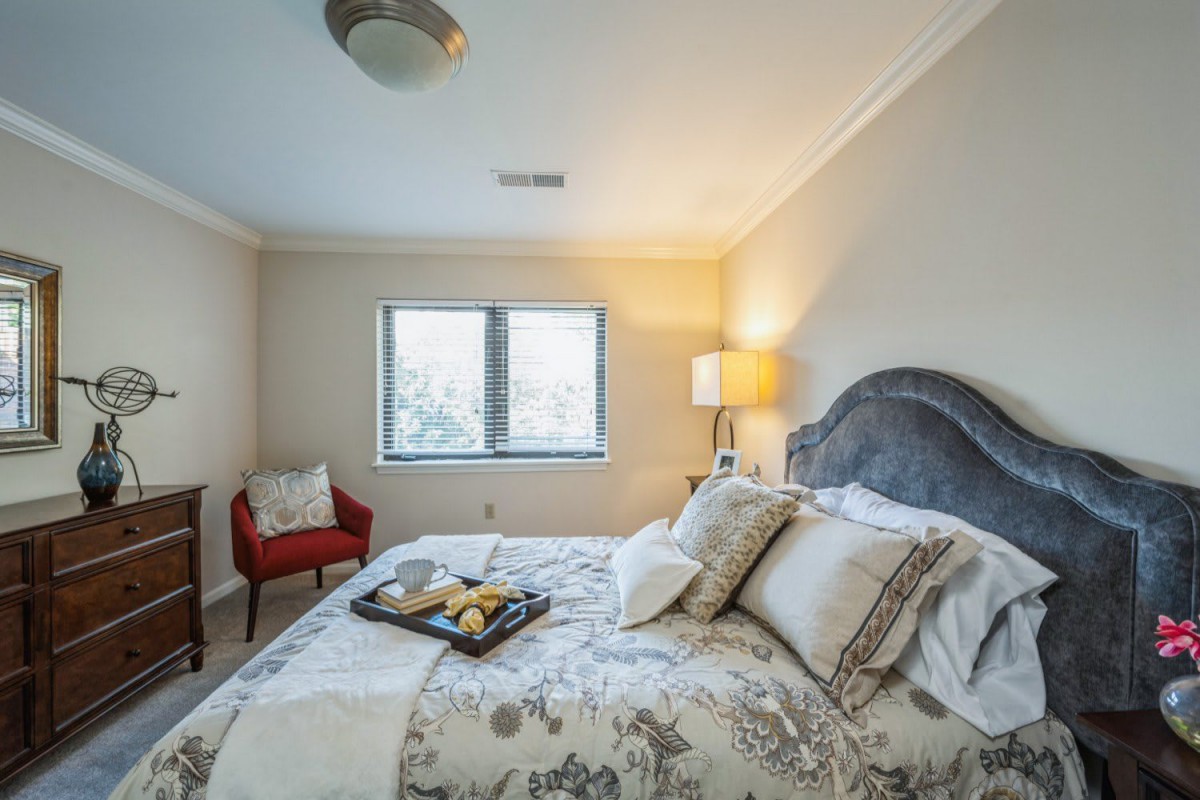 A cozy bedroom with a neatly made bed, decorative pillows, and a tray on the bed. A red chair and lamp are positioned by the window.