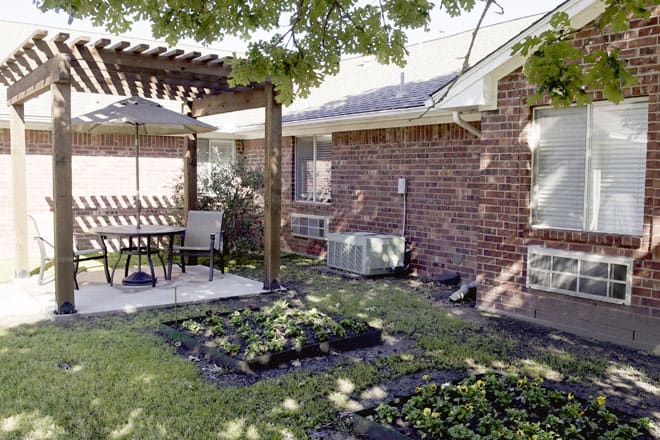 A cozy backyard with a pergola-covered patio, featuring outdoor seating and a small garden alongside a brick house.