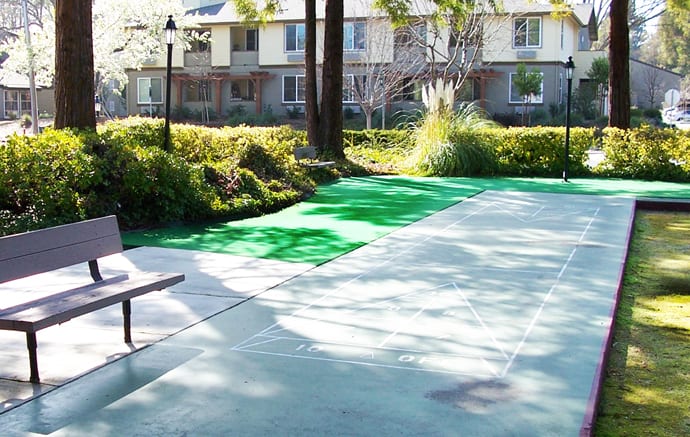 A sunny outdoor pathway near a residential area, surrounded by greenery and benches.