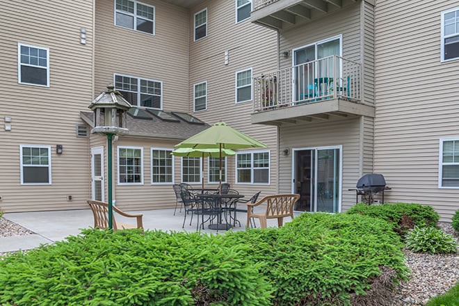 A cozy apartment patio with outdoor seating and a green umbrella, surrounded by lush greenery.