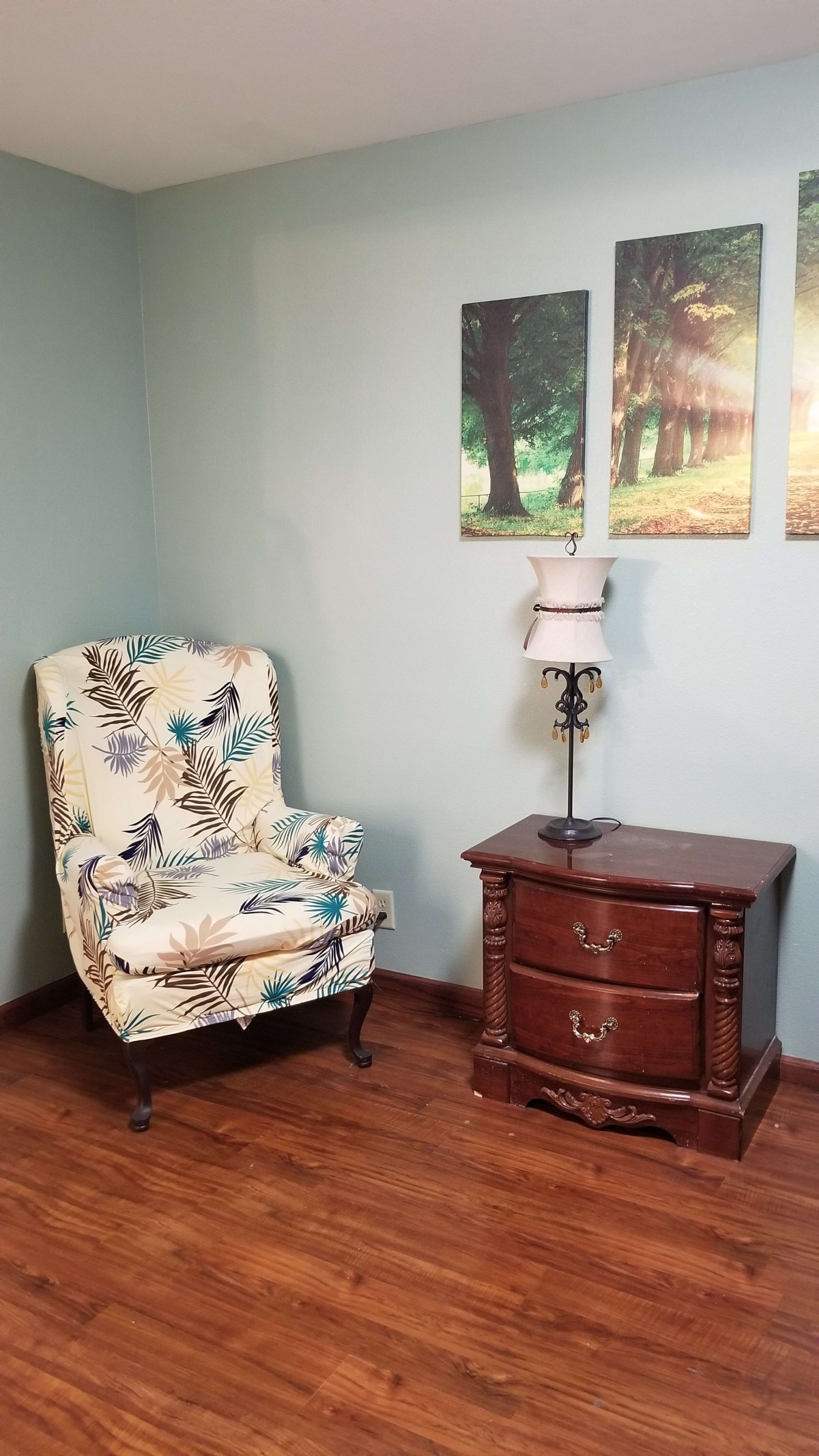 A cozy corner featuring a patterned armchair, a wooden side table with a lamp, and forest-themed wall art.