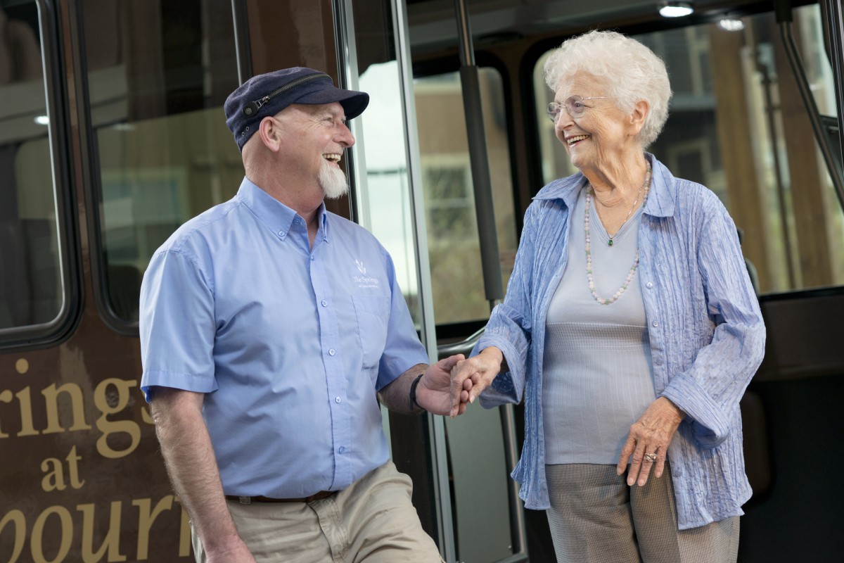 Two elderly individuals smiling and holding hands while standing outside.