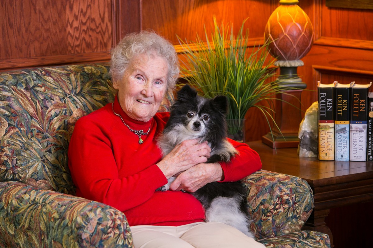 An elderly woman sitting on a chair, smiling and holding a small black and white dog in a cozy living room setting.