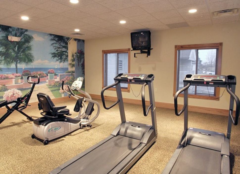 A gym with two treadmills and a stationary bike, featuring large windows and a wall-mounted TV.