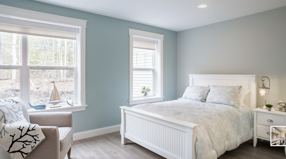 A minimalist bedroom featuring soft blue walls, a white bed frame, and ample natural light from two windows.