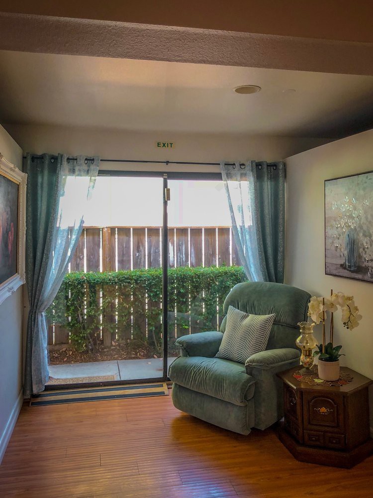 A cozy living room with a comfortable armchair facing a sliding glass door, adorned with sheer curtains and greenery.
