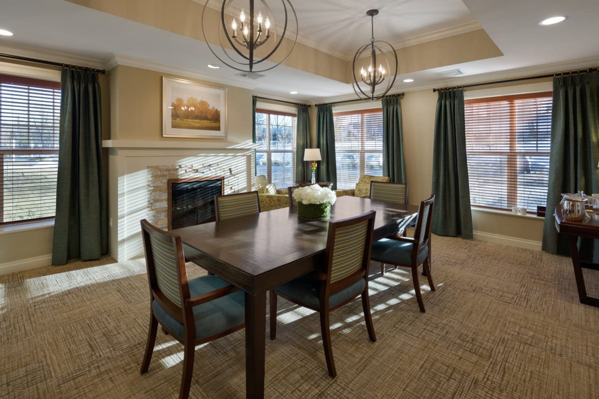 A modern dining room with a wooden table, upholstered chairs, and stylish chandeliers, featuring a fireplace and large windows with curtains.