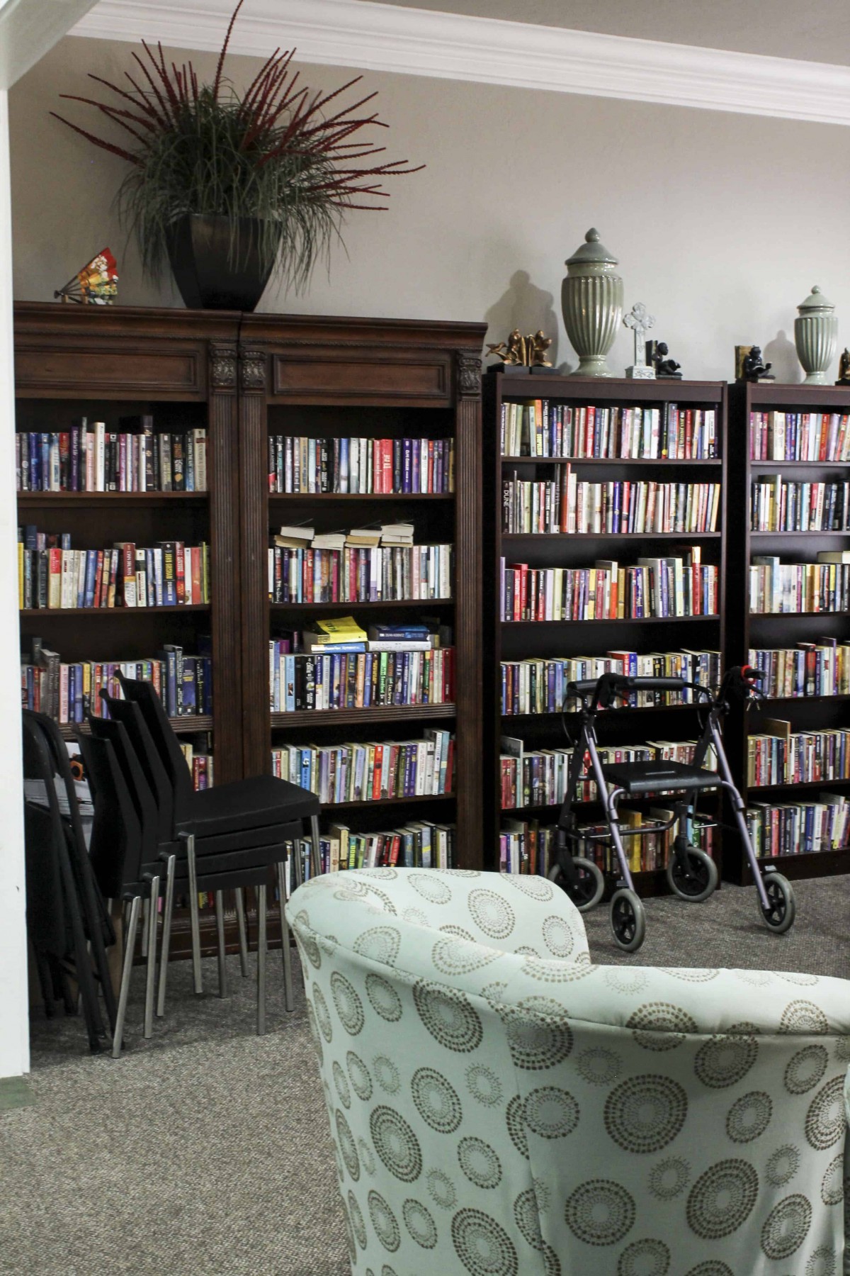 A cozy library setting with bookshelves filled with books, chairs arranged in a corner, and a walker nearby.