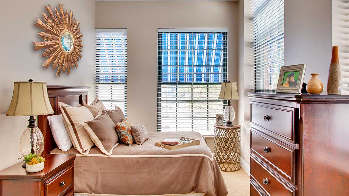 A cozy bedroom with warm tones, featuring a neatly made bed, two large windows with blinds, a bedside lamp on a decorative table, and a wooden dresser.