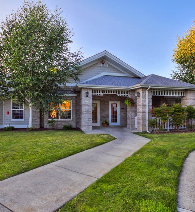A charming single-story house with a well-manicured front lawn and trees, featuring a welcoming pathway leading to the entrance.