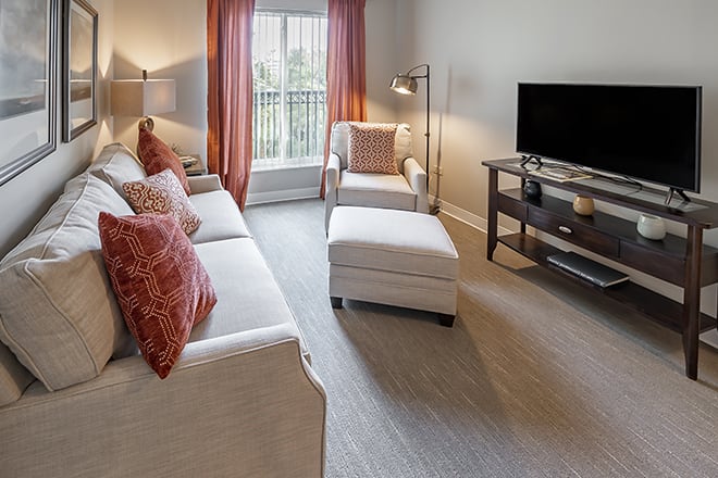 A cozy living room with a sofa, armchair, and ottoman arranged around a television, featuring warm curtains and soft lighting.