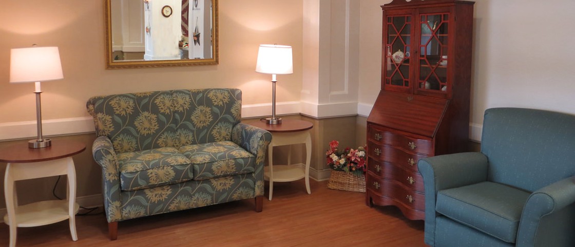 A cozy living room corner featuring a floral-patterned sofa, an elegant side table with a lamp, a decorative wooden cabinet, and a mirror on the wall.