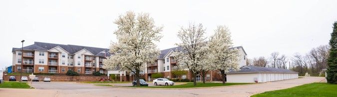 An apartment building surrounded by blooming trees with white flowers in the spring, featuring parked cars nearby.