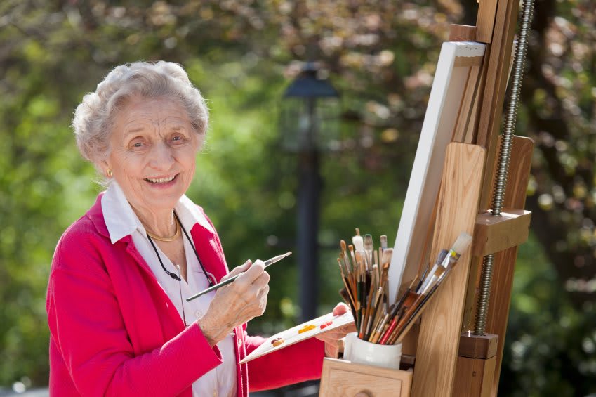 An elderly woman painting on a canvas outdoors, surrounded by art supplies and an easel.