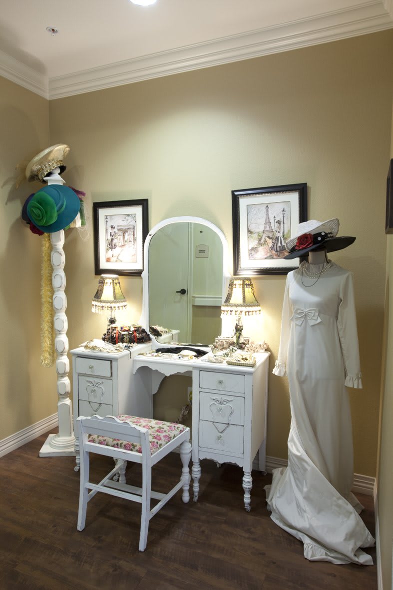 A vintage-themed room with a white vanity, a mannequin wearing a wedding dress, and various hats displayed.