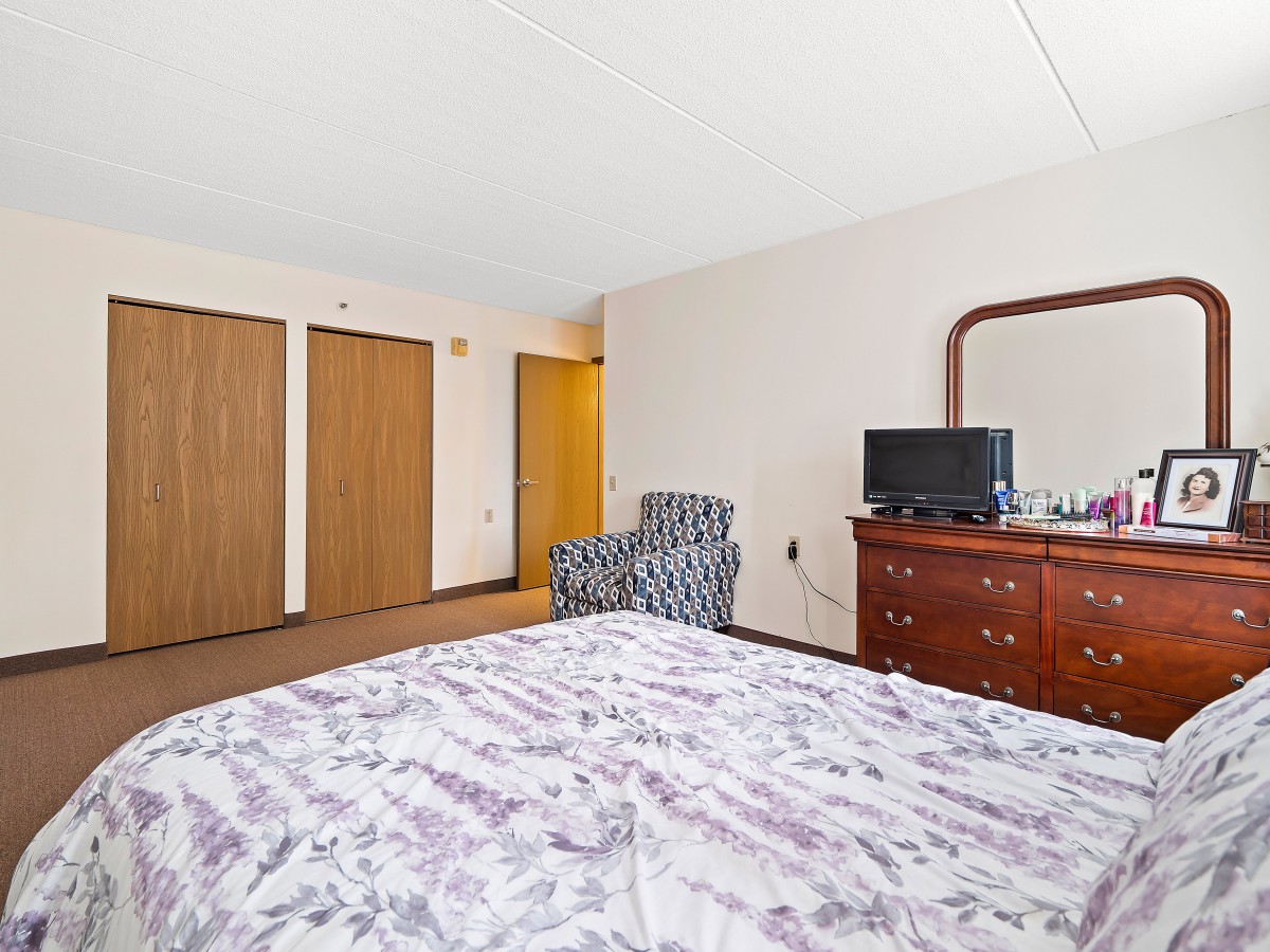 A bedroom with a bed, dresser, television, and patterned chair.