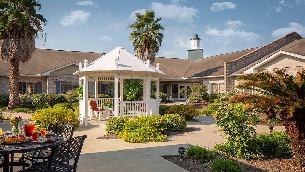 A serene garden featuring a white gazebo surrounded by lush greenery and a well-maintained pathway, set against the backdrop of a large building with a tower.