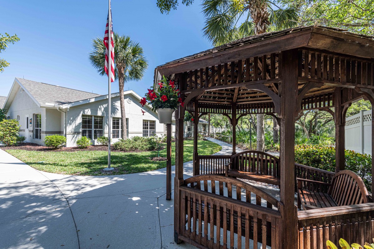 A wooden gazebo in a landscaped garden with an American flag and a nearby building.