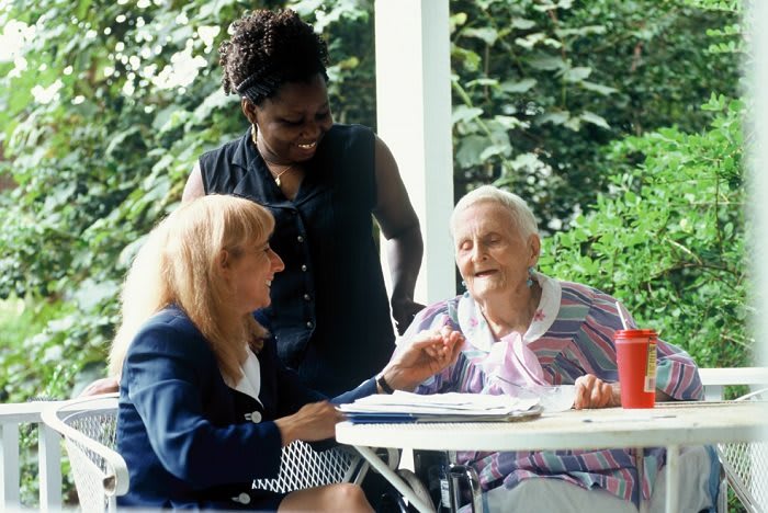 Two caregivers talking and smiling with an elderly woman sitting at a table outdoors.