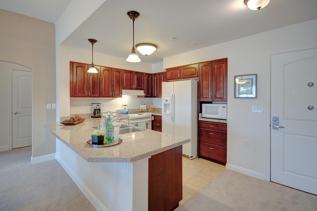 A modern kitchen with wooden cabinets and white countertops, featuring appliances and pendant lighting.