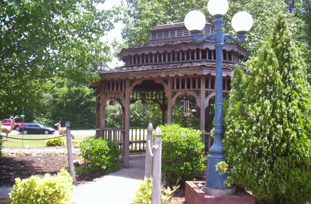 A wooden gazebo in a park surrounded by lush greenery and a lamp post.