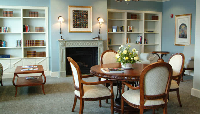 A cozy living room with a round table, chairs, and a fireplace, surrounded by bookshelves and decorated with flowers.