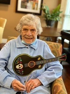 An elderly woman smiling while holding a decorative guitar indoors.