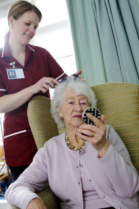 An elderly woman sitting in a chair uses a mirror as a caregiver brushes her hair.