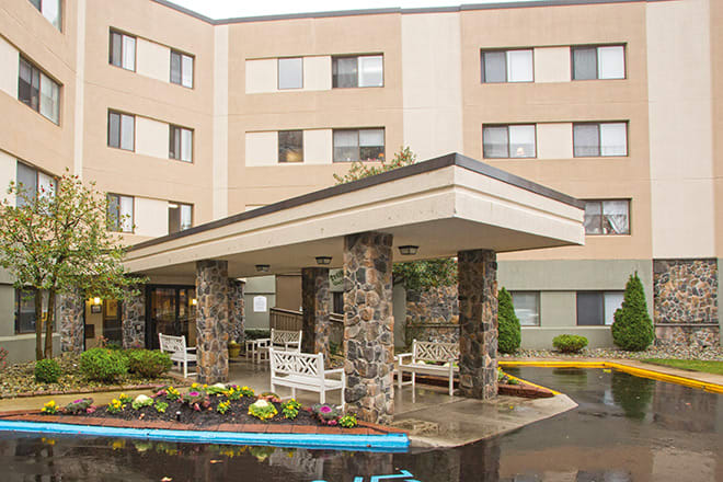 The image shows the entrance of a multi-story building with stone columns and benches, surrounded by landscaped areas.