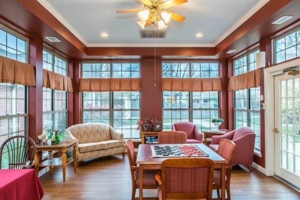 A cozy living room with abundant natural light, featuring a mix of comfortable seating and a table set, highlighted by large windows and warm wooden flooring.