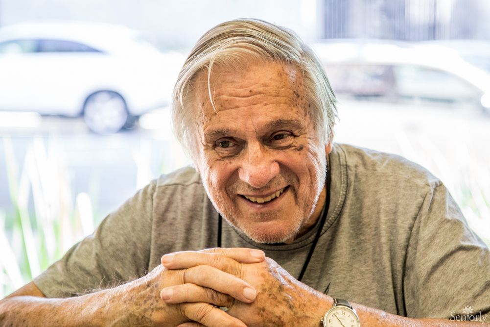 An elderly man smiling while sitting indoors with hands clasped.