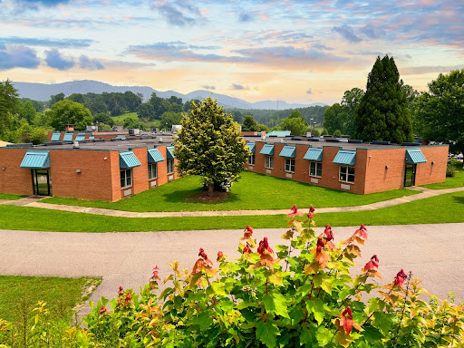A scenic view of a school building surrounded by lush greenery and mountains under a cloudy sky.