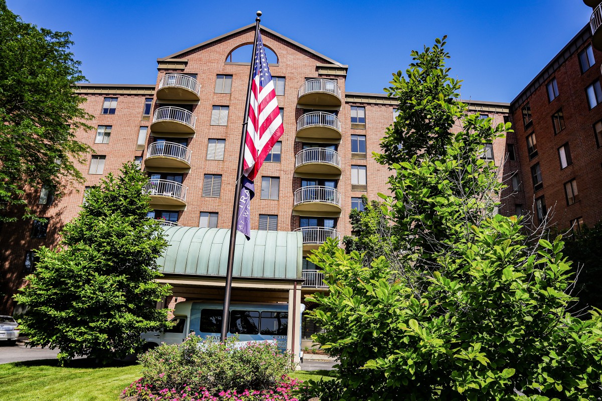 A multi-story residential building with balconies, surrounded by trees, featuring a prominently displayed American flag at the entrance.