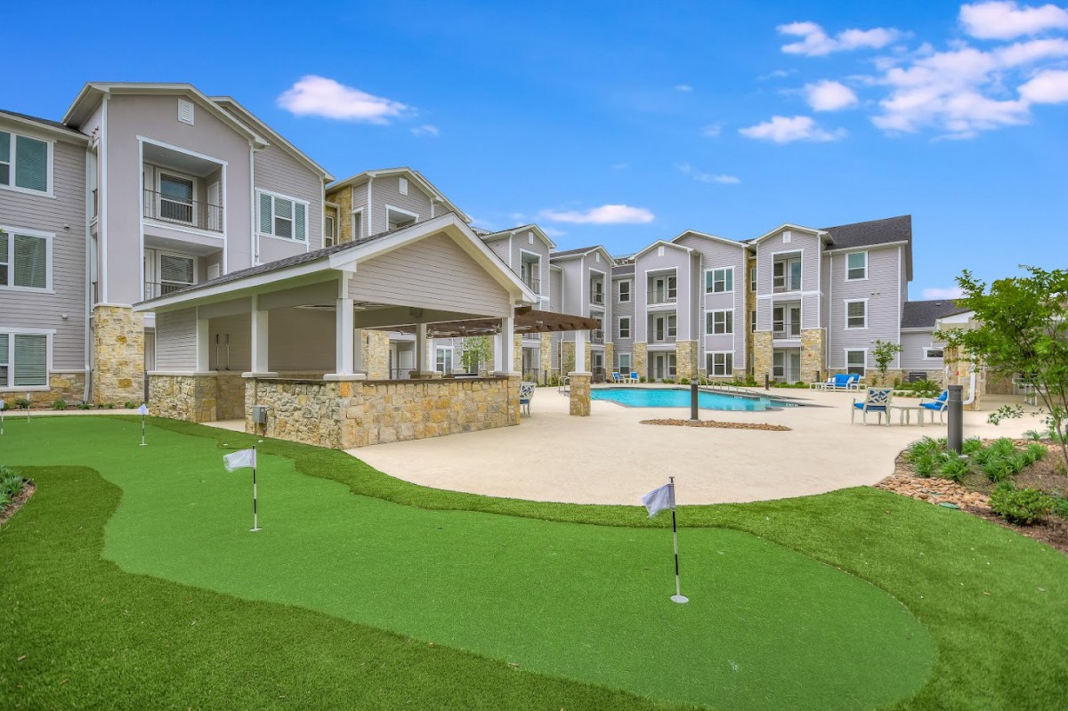 A modern apartment complex features outdoor amenities including a swimming pool and a golf putting green on a sunny day.