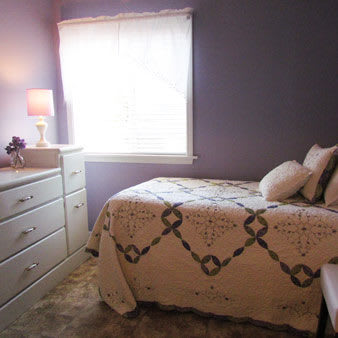 A cozy bedroom with purple walls, a neatly made bed, and a dresser beside a window letting in natural light.