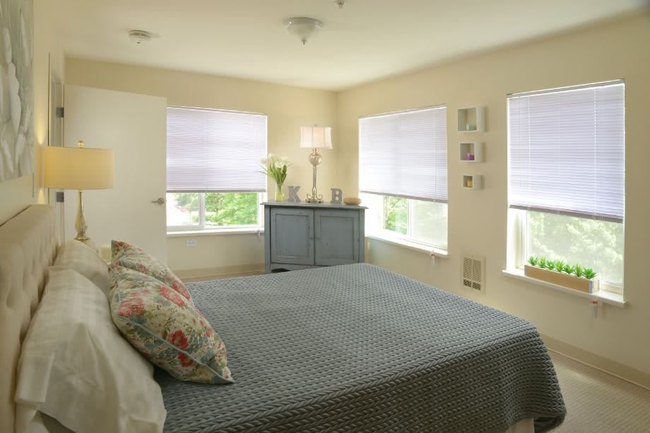 A cozy bedroom with ample natural light, featuring a bed, bedside table, and large windows.