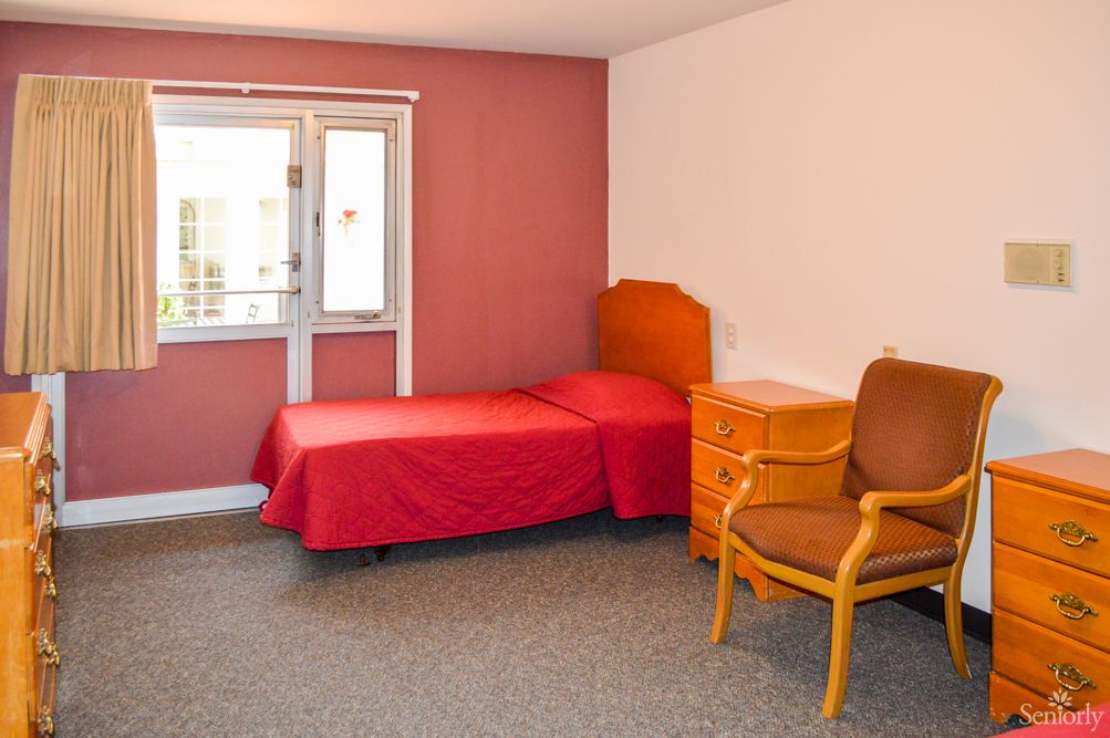 A simple bedroom with a single red-bedded bed, wooden nightstand, and a chair next to a window.