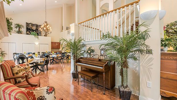 A spacious living area with a piano and dining space, accented by potted plants and a wooden staircase.