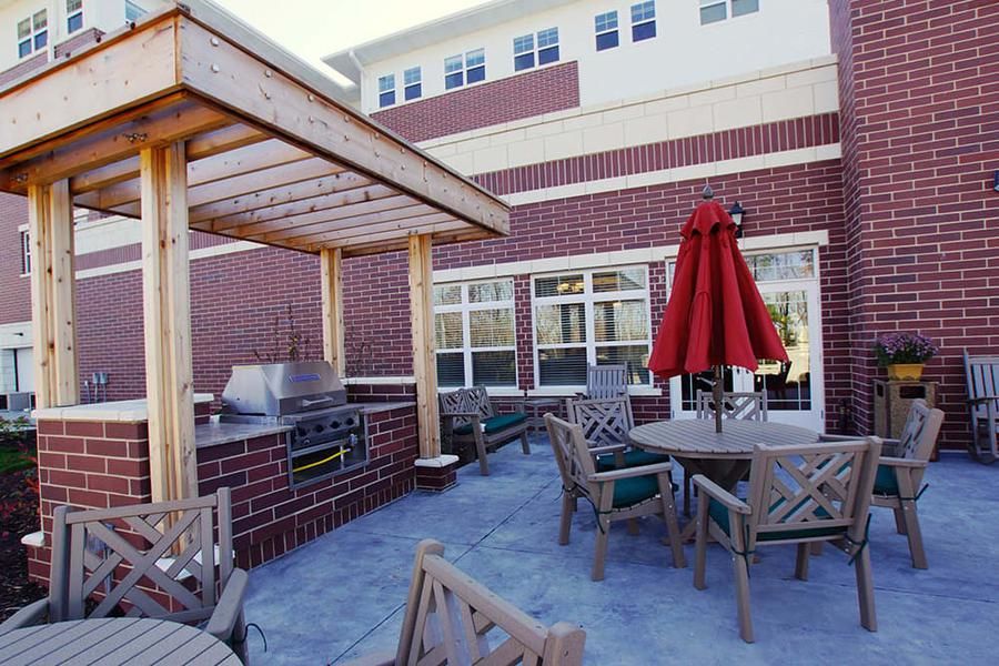 An outdoor patio area with tables, chairs, a red umbrella, and a grill under a wooden pergola against a brick building.